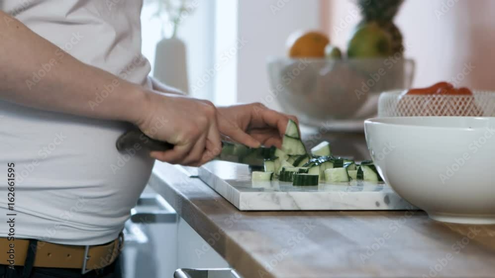 A pregnant woman stands in her kitchen cutting a cucumber. A closeup shot of her hands while chopping. A pregnant woman stands in her kitchen cutting a cucumber. A closeup shot of her hands while chopping.