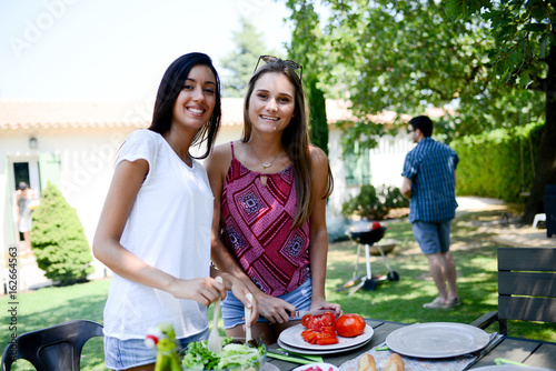 two beautiful young woman in garden preparing tomato salad for barbecue party during summer holiday