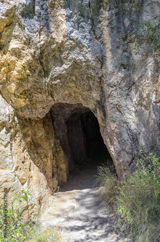 Entrance to gallery of a rocky mountain. Hiking trail near to Montanejos town dam, in the province of Castellon (Valencian Country - Spain). At the shadow of the trees. Darkness at the background.