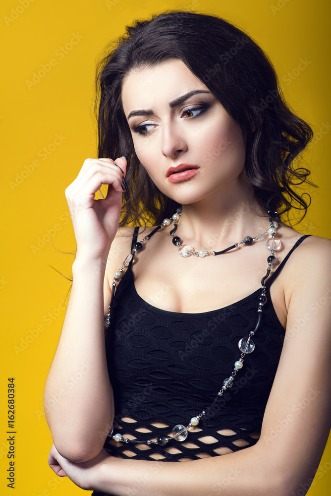 Obraz premium Portrait of a young beautiful dark haired concerned woman wearing black net top and glass beads looking worried and upset. Isolated on yellow background. Studio shot