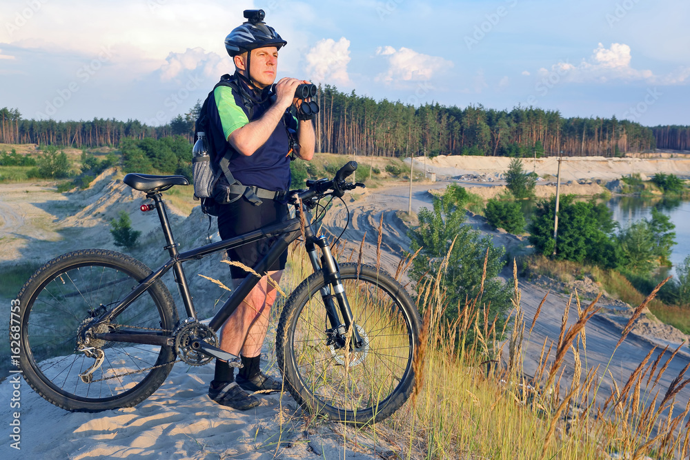 The cyclist with the Bicycle and binoculars in hand stands on a sandy hill in the sunlight.
