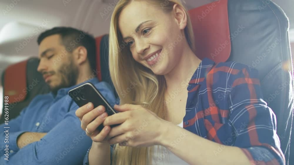 On Board of Commercial Airplane Beautiful Young Blonde Uses Smartphone while Her Hispanic Male Neighbor Sleeps. Shot on RED EPIC-W 8K Helium Cinema Camera. On Board of Commercial Airplane Beautiful Young Blonde Uses Smartphone while Her Hispanic Male Neighbor Sleeps. Shot on RED EPIC-W 8K Helium Cinema Camera.