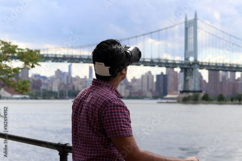 Young handsome man wearing virtual reality goggles headset while standing infont of waterfront bridge and urban city skyline photo