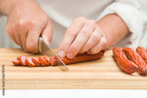 Chef's hands with knife cutting smoke-dried sausages on the wooden board. Preparation for cooking. Healthy eating and lifestyle.