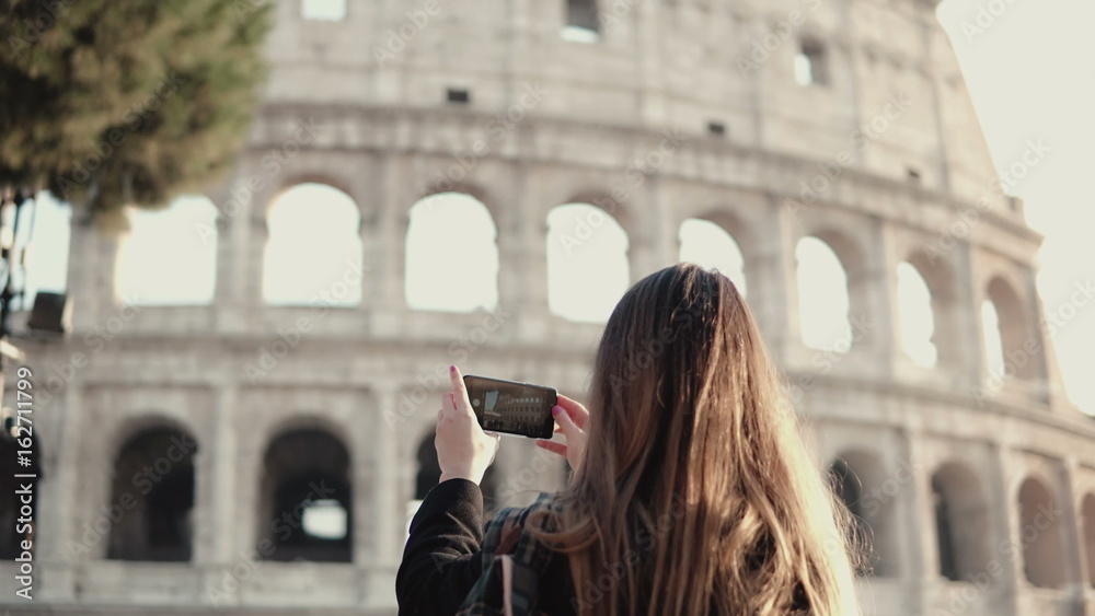 Young brunette tourist exploring the Colosseum in Rome, Italy. Woman takes the photo of sight, uses smartphone.