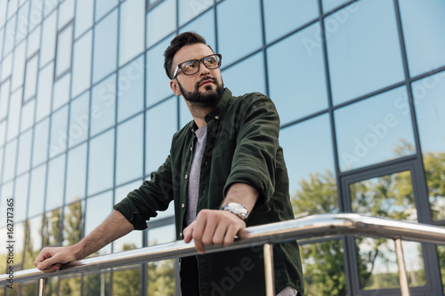 Handsome bearded man in eyeglasses leaning at railing at looking away