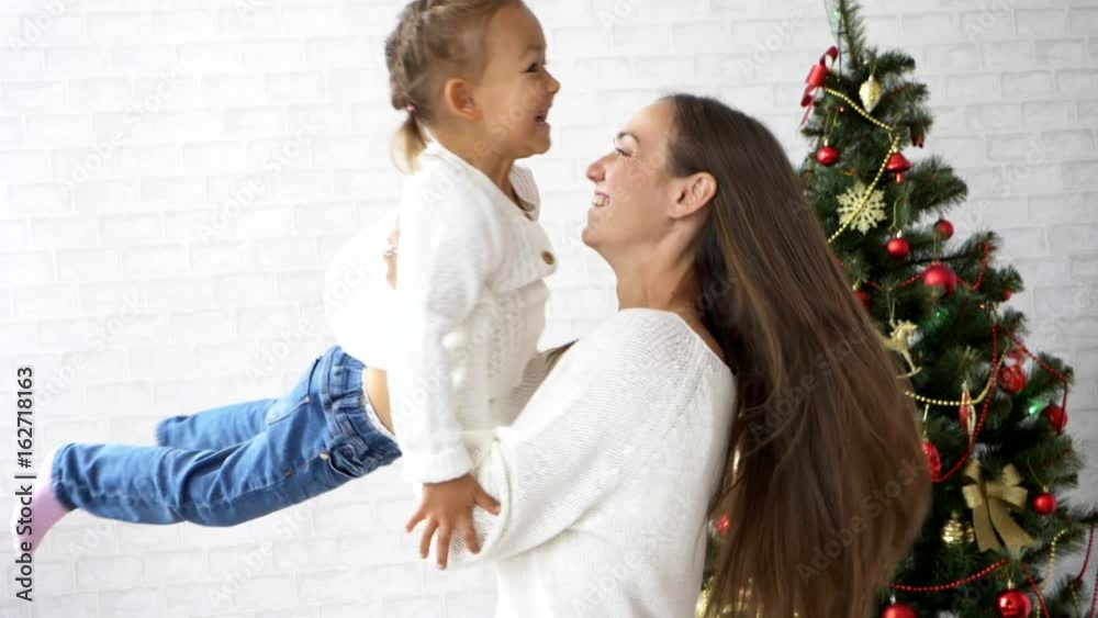 Young mother turns her little daughter with Christmas tree on the background Young mother turns her little daughter with Christmas tree on the background