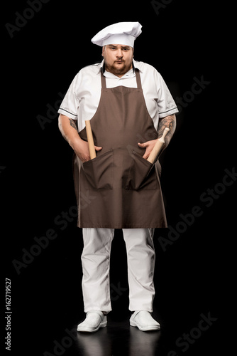 Professional young chef in hat standing with rolling pins n pockets of apron and looking at camera © LIGHTFIELD STUDIOS