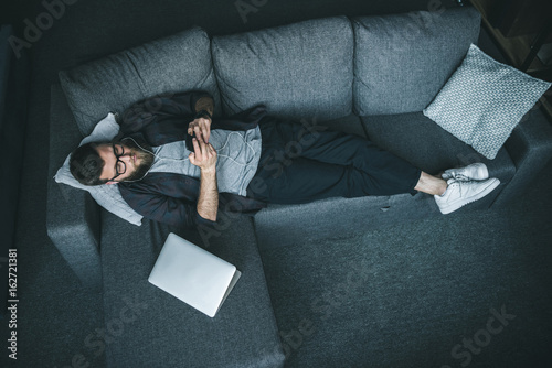 High angle view of bearded young man in eyeglasses and earphones using smartphone while lying on sofa