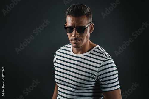 serious trendy man in striped t-shirt and sunglasses looking at camera, isolated on grey