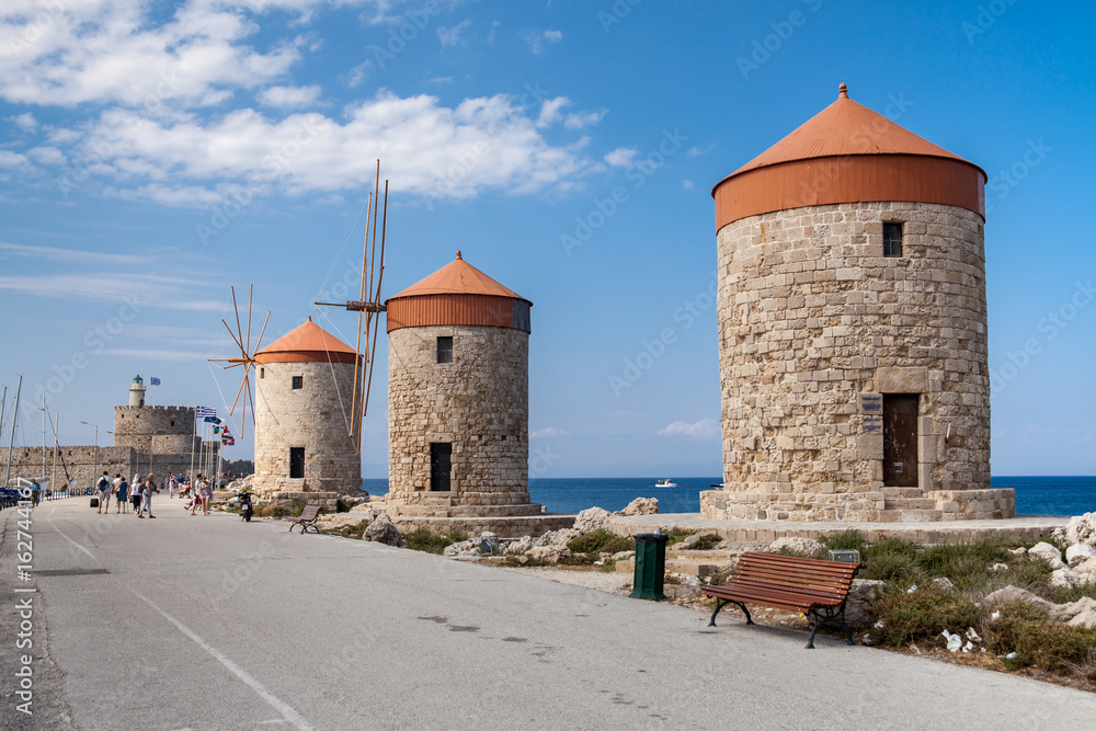 Windmill Papagiorg whit Vati in Rhodes harbor. Old defensive stands and windmills. Wharf harbors, boats and sailing ships. Historic pier and beach.