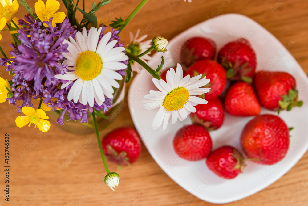 Bouquet of wildflowers and lilacs in a glass jar, next to a white dish with fresh strawberries on wooden table, top view, selective focus