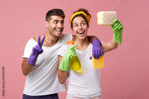 Funny couple having fun during doing housework and cleaning: smiling man hugging his wife who is making selfie with sponge. Cheerful male and female using sponge like smartphone making selfie together