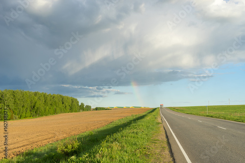 Fanciful cloudscape and rainbow over farm buildings with vanishing highway at sunset. Silkovo, Kaluzhsky region, Russia. 
