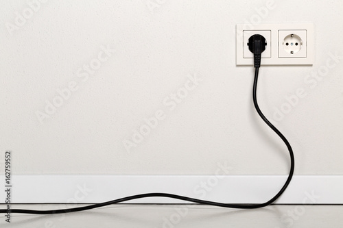 Black power cord cable plugged into european wall outlet on white plaster wall photo