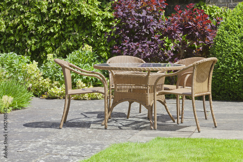Fototapeta Naklejka Na Ścianę i Meble -  Rattan dining chairs and table in a garden patio surrounded by plants and flowers, on a sunny summer day .