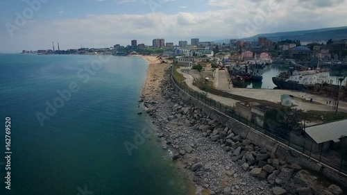 City on Shore. Bouldings on Hill. Boat Staying in Port. Road Along Sealine. Resort Houses on Beach. Blue Water. Cloudy Summer Day photo