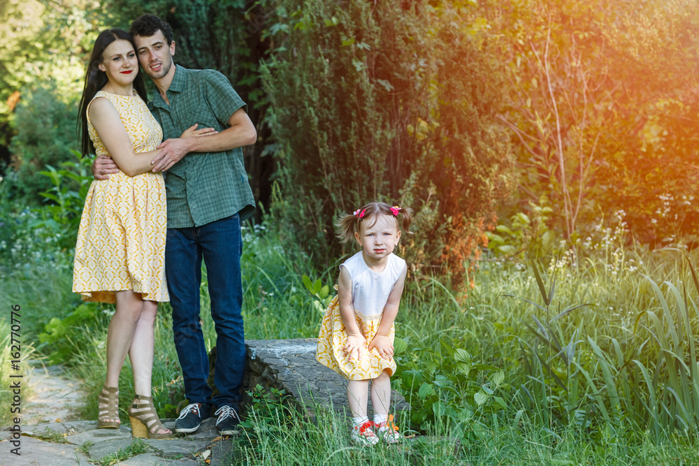 Fototapeta premium In foreground a girl is standing with his hands on his knees. In background parents embracing each other looking at her.