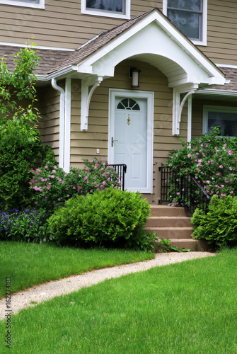 House entrance with white front door and stairs at summer. White front door with roofed porch of beige sided house. Seasonal plants surround the stairs and green grass lawn with sidewalk in forefront.