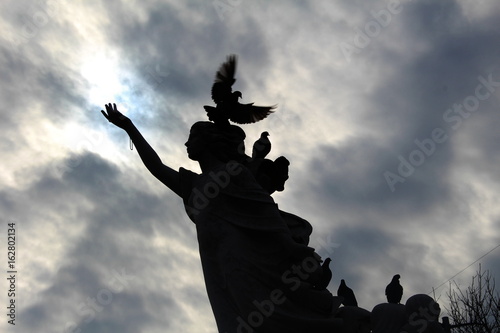 Statue of girl silhoetted by pigeons with a cloudy sun behind -making her look like an angel photo