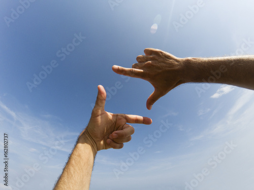 Two hands form with a fingers a frame in front of a blue sky with clouds and reflection