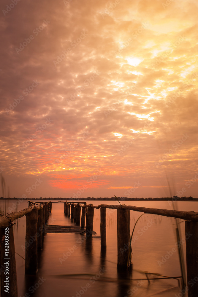 long exposure of wooden old bridge into the lake on the background of worm sky sunset.
