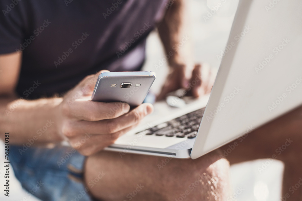 Young man using smartphone and laptop in the city. Close up of cheerful adult using gadgets on a street Young man using smartphone and laptop in the city. Close up of cheerful adult using gadgets on a street
