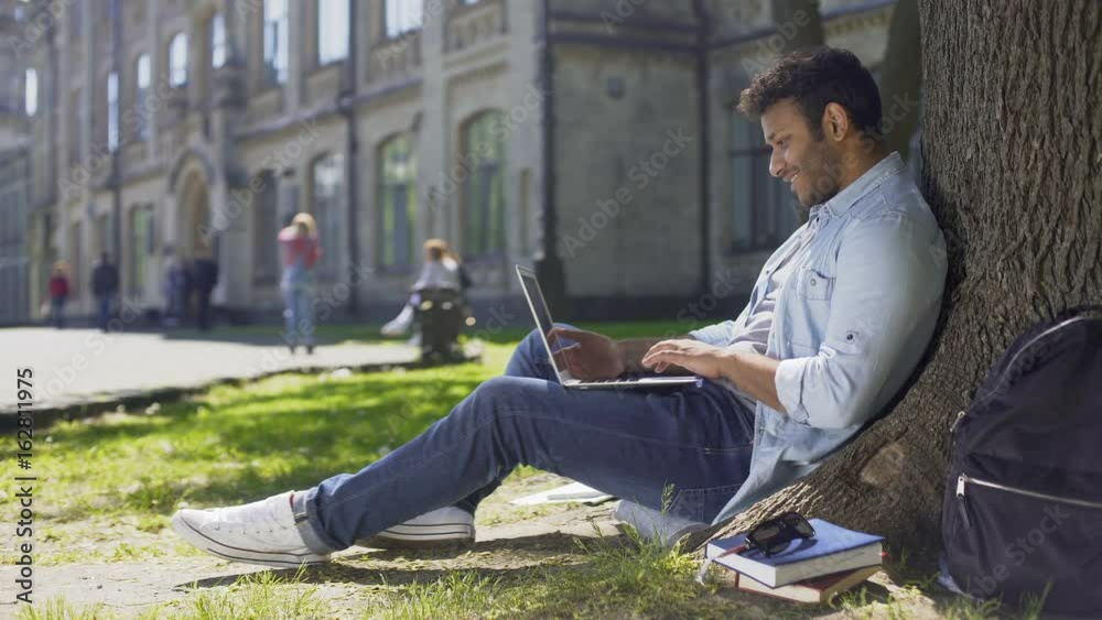 Young mixed-race guy using laptop under tree, surprised reaction, good news Young mixed-race guy using laptop under tree, surprised reaction, good news