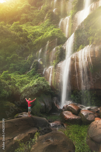 Young strong man traveler enjoy spectacular morning view, beautiful Waterfall cascade hidden in tropical jungle on the background green tree forest nature and mountain