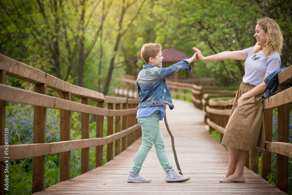 Obraz premium Portrait of young blond woman standing on a wooden bridge and giving five to her cute son in the park on a sunny summer day. Lifestyle. Outdoor.