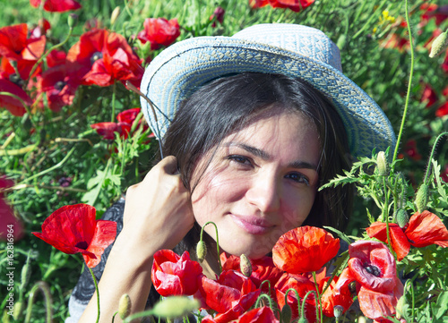Portrait of beautiful brunette in a summer hat against a background of red poppies in the height of summer.Beautiful woman enjoying the bright red wild flowers, harmony concept. photo
