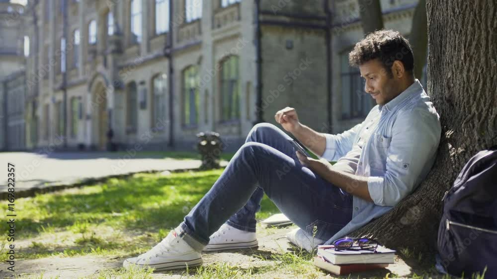 Male sitting under tree, using mobile phone, reacting to newsfeed, confusion Male sitting under tree, using mobile phone, reacting to newsfeed, confusion