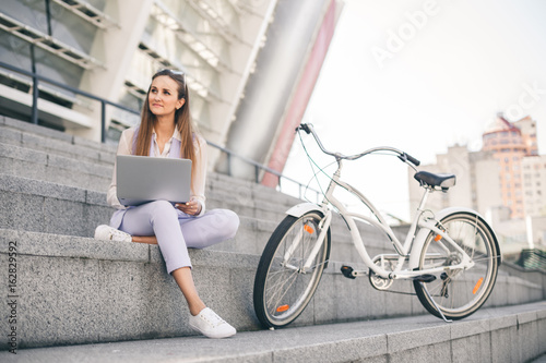 Beautiful woman office worker at lunch break working on laptop. A young woman is sitting on the steps, next to the bike.