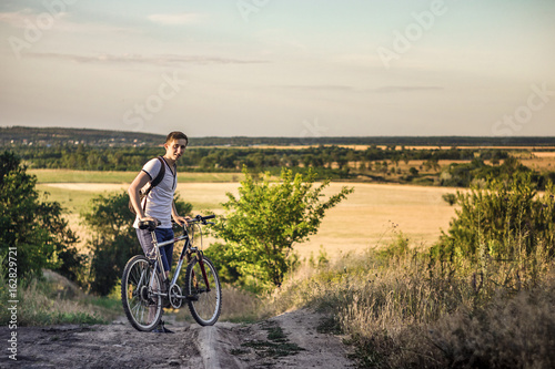 A tourist man stands holding a bicycle in front of an opening landscape under a blue sky