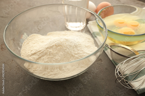 Glass bowl with flour and ingredients for dough on grey table