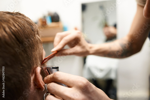 Close up shot of young European male customer getting his hair cut by professional skillful hairdresser who is using comb and electric razor. Beauty industry, haircare and hairdressing. Film effect