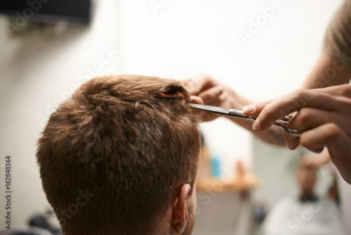 Rear view of guy sitting at barbershop, having his hair cut by unrecognizable hairdresser. Master cutting male client's hair using comb and scissors at professional hairdressing salon. Selective focus