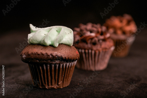 Three chocolate cupcakes garnished with chocolate chips and chocolate and mint frosting on lined up across the frame, on black background photo