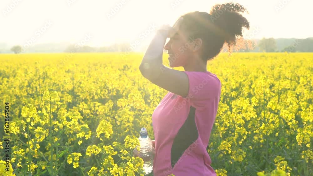 4K video clip of beautiful healthy mixed race African American girl teenager female young woman running or jogging and drinking a bottle of water in field of yellow flowers 4K video clip of beautiful healthy mixed race African American girl teenager female young woman running or jogging and drinking a bottle of water in field of yellow flowers