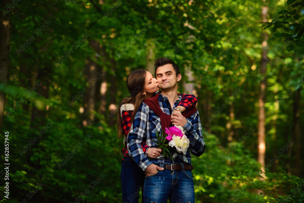 Stylish young couple in shirts and jeans while walking in the forest. A charming girl kisses on the cheek her handsome boyfriend. Stylish young couple in shirts and jeans while walking in the forest. A charming girl kisses on the cheek her handsome boyfriend.