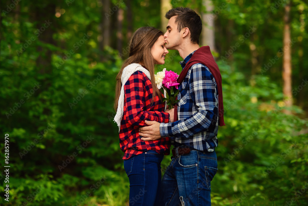 Fototapeta premium Stylish young couple in shirts and jeans while walking in the forest. A handsome boyfriend kisses his beautiful charming girlfriend on the forehead.