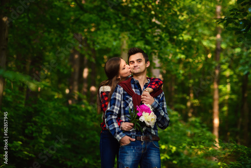 Stylish young couple in shirts and jeans while walking in the forest. A charming girl kisses on the cheek her handsome boyfriend.