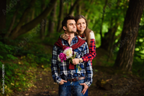 Stylish young couple in shirts and jeans while walking in the forest. Beautiful charming girl hugs her handsome boyfriend.