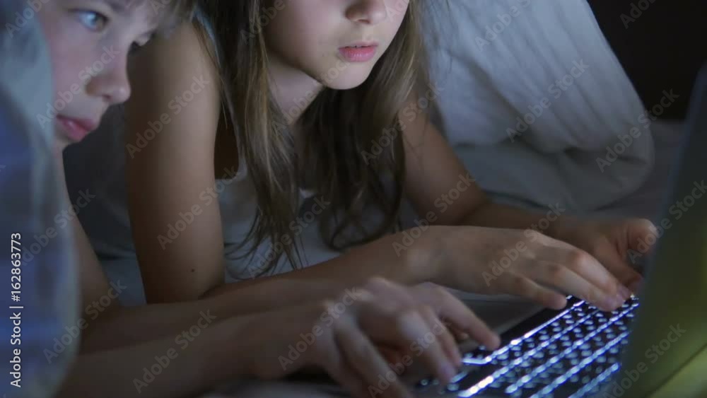 A boy and a girl are playing on the computer and lying on the bed under the blanket at night.