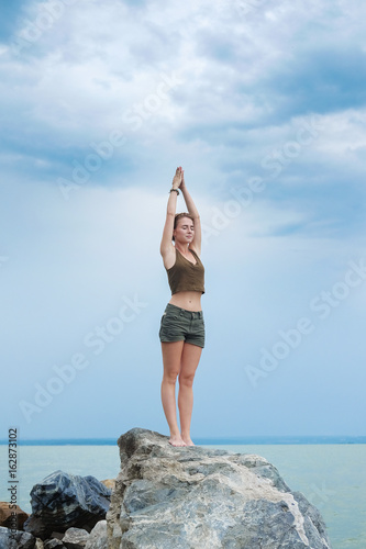 Girl practicing yoga on the rocks against the blue sky and the azure sea. Woman raises her arms to the sky in the pose of namaste.