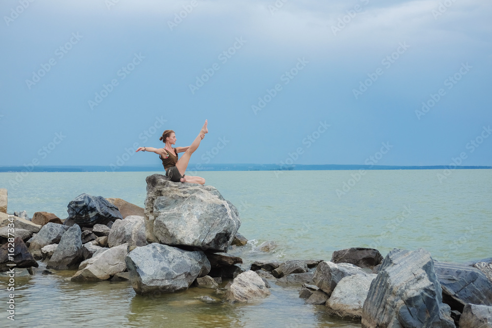 Girl practicing yoga on the rocks against the blue sky and the azure sea. Relaxation and Stretching. Young slim sporty woman do yoga outdoors.
