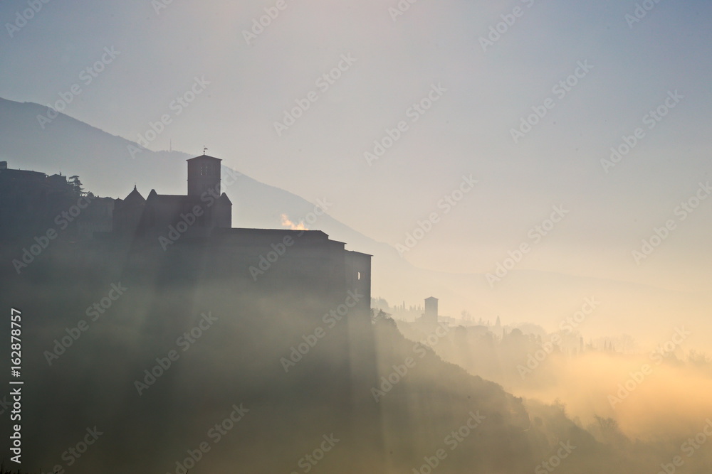 A view of a silhouette of St.Francis church in Assisi in the middle of mist, with sun rays projecting shadows and beautiful, warm sunset colors