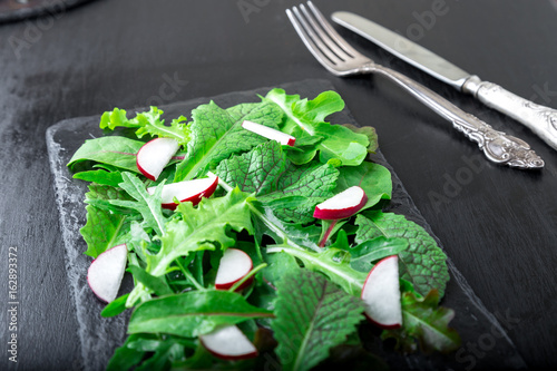 Mixed salad leaves with radish on black slate plate. Closae up.