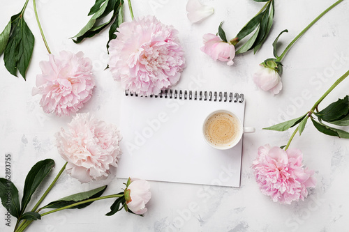 Morning coffee cup for breakfast, empty notebook and pink peony flowers on white stone table top view in flat lay style. Woman working desk.
