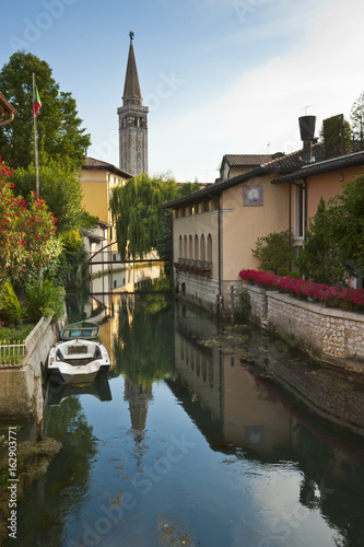 The tower bell of the cathedral of St. Nicolò is reflected on Livenza river in Sacile. Italy